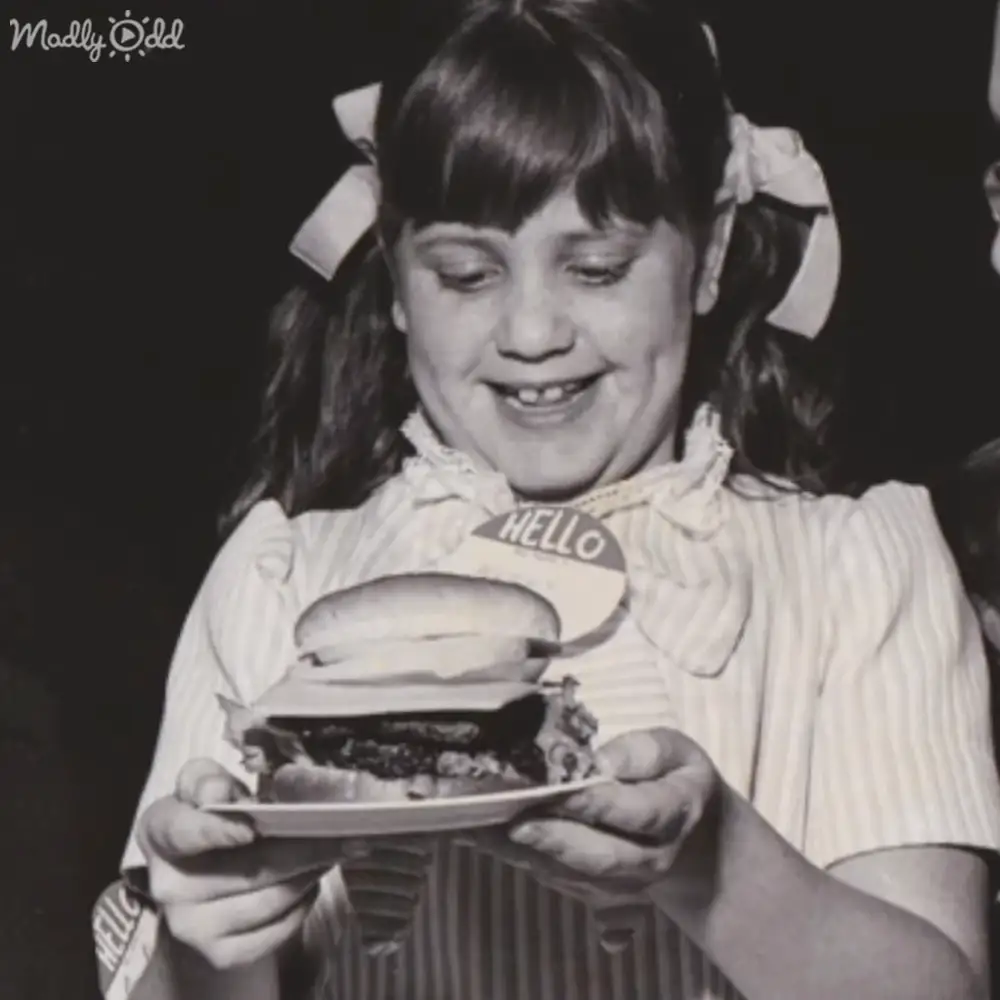 A child wearing a vintage-style white puffed-sleeve blouse or dress with ruffled details holds a large hamburger indoors.