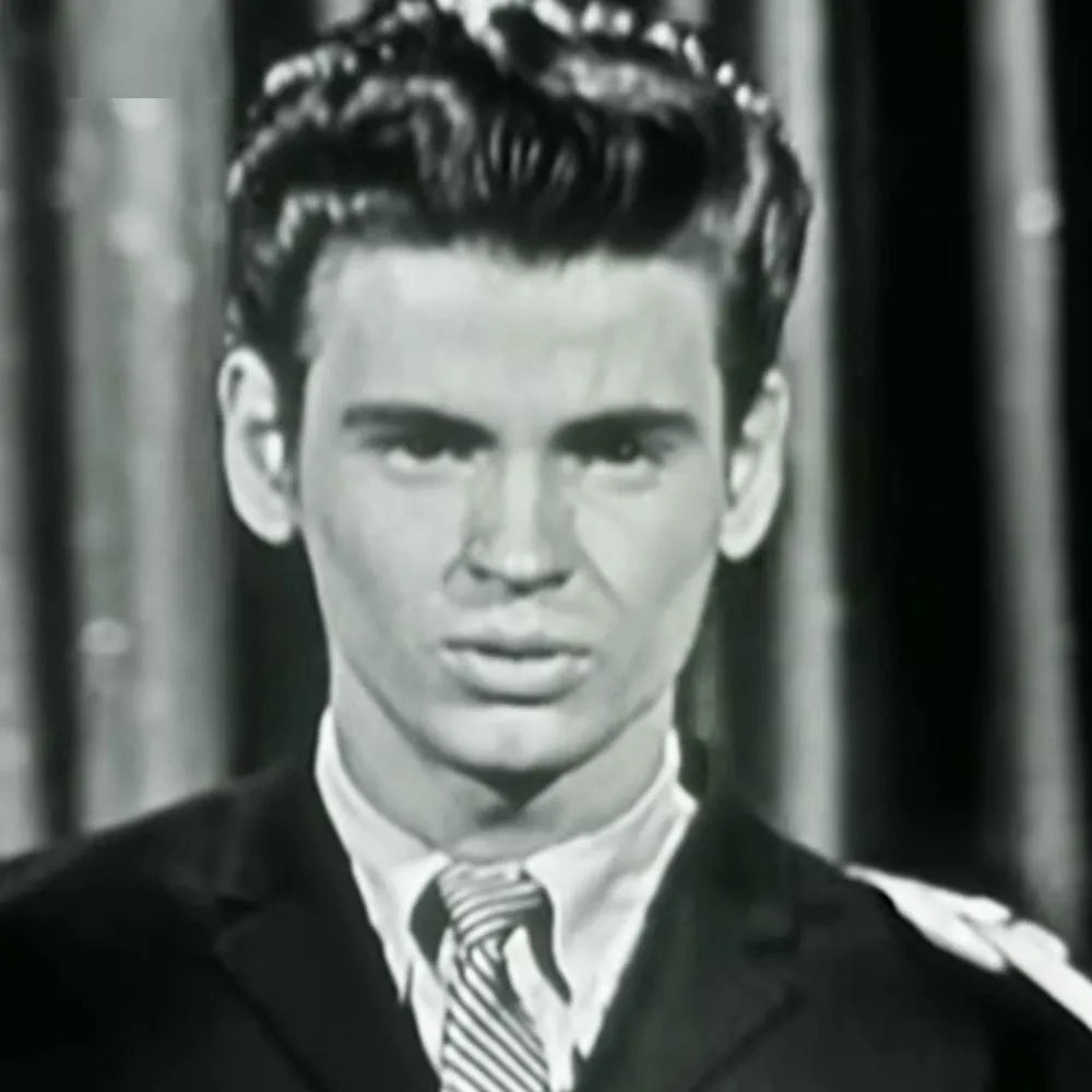 A young adult male in a tailored suit jacket with a dark, collared shirt, standing indoors against vertical blinds.