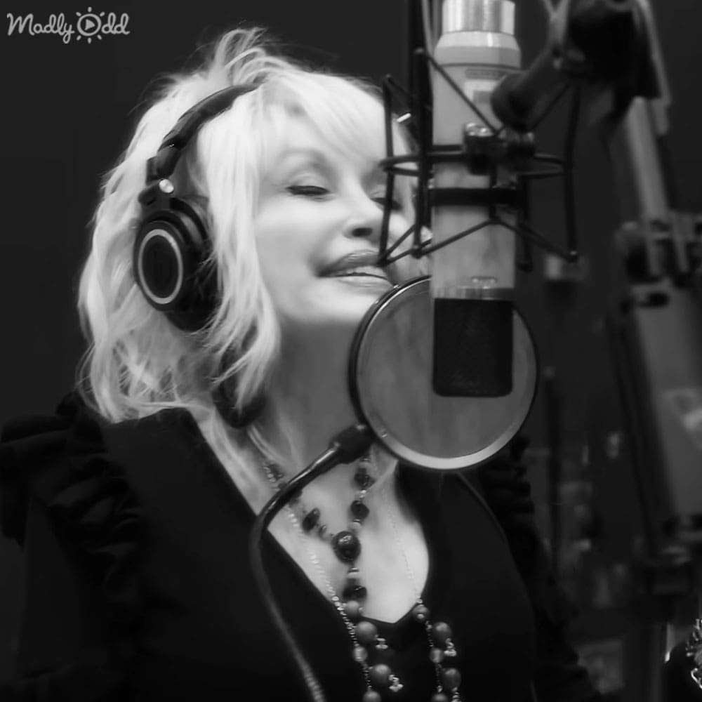 A middle-aged adult female in a recording studio wearing a loose-fitting, elegant black top with ruffled sleeves and large black headphones around her neck.