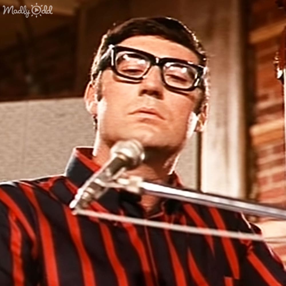 A middle-aged adult male wearing a red and black striped shirt with collared long sleeves, large dark-framed glasses, standing indoors near a microphone stand.