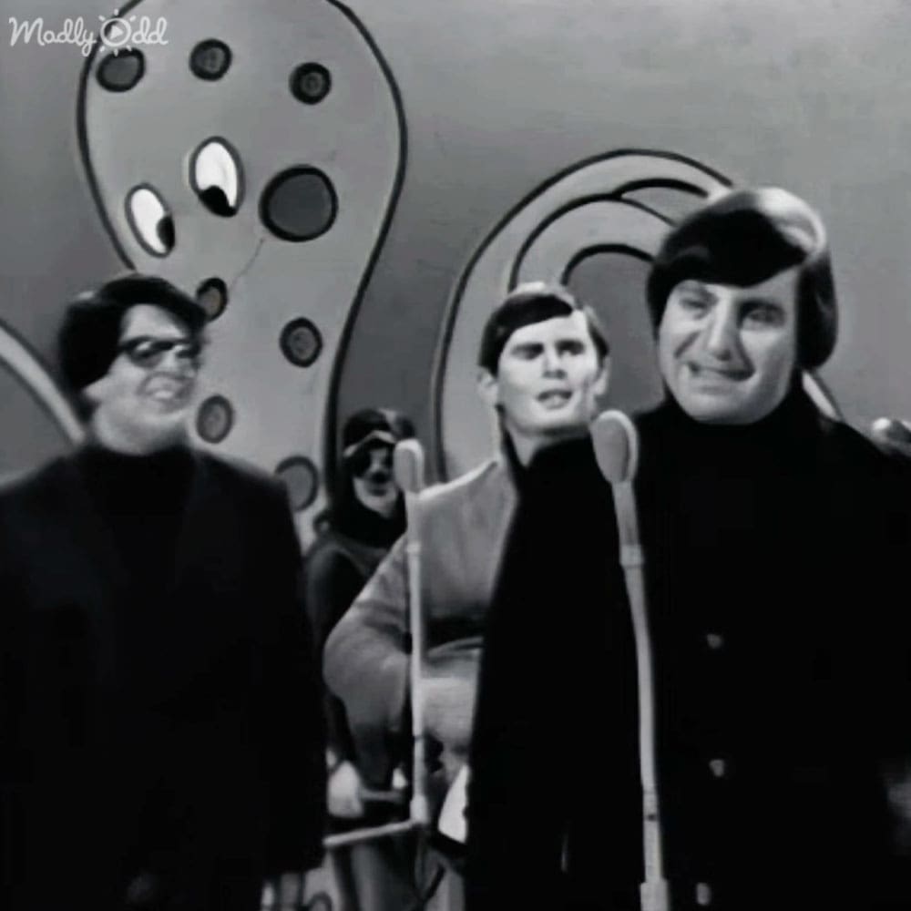 A mid-20th century musical performance scene with three adult males wearing dark, textured jackets and collared shirts on a stage with futuristic light gray abstract shapes in the background.