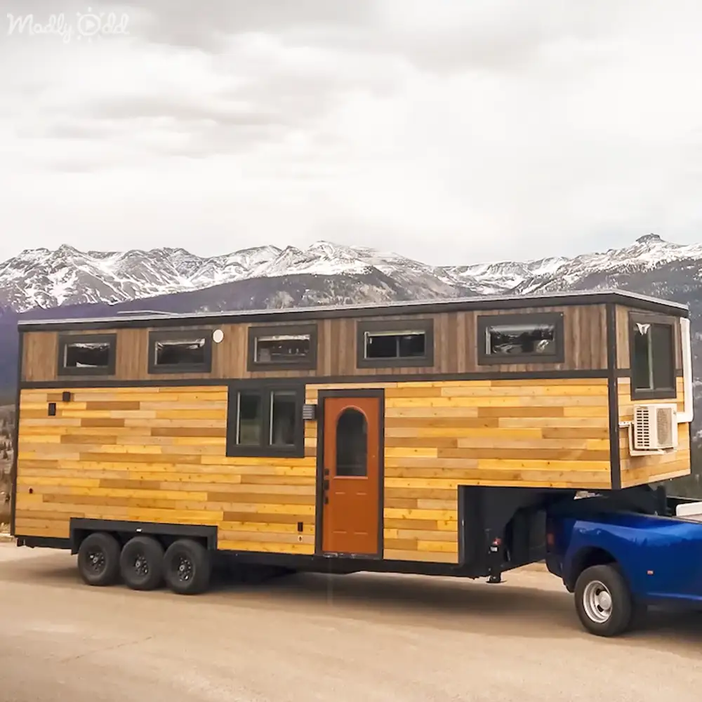 A wooden tiny house trailer being towed by a blue pickup truck, set against snow-capped mountains under an overcast sky during winter or early spring.