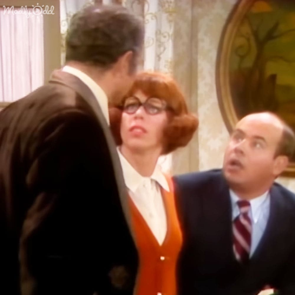 An adult female wearing an orange dress with a white collar and buttons down the front, standing indoors near two men dressed in dark suits under ornate curtains and framed artwork on light-colored walls.