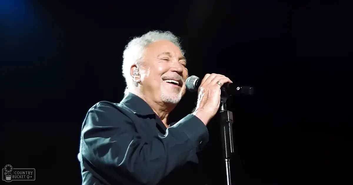 A middle-aged adult male singer performing on stage in a relaxed, dark blue or black collared shirt with an earpiece and ring, illuminated by focused lighting against a dark background.