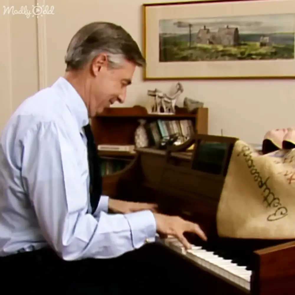 A middle-aged to older adult male in a light blue dress shirt with long sleeves and a dark tie, playing the piano indoors, surrounded by books, decorative items, and a framed landscape painting.