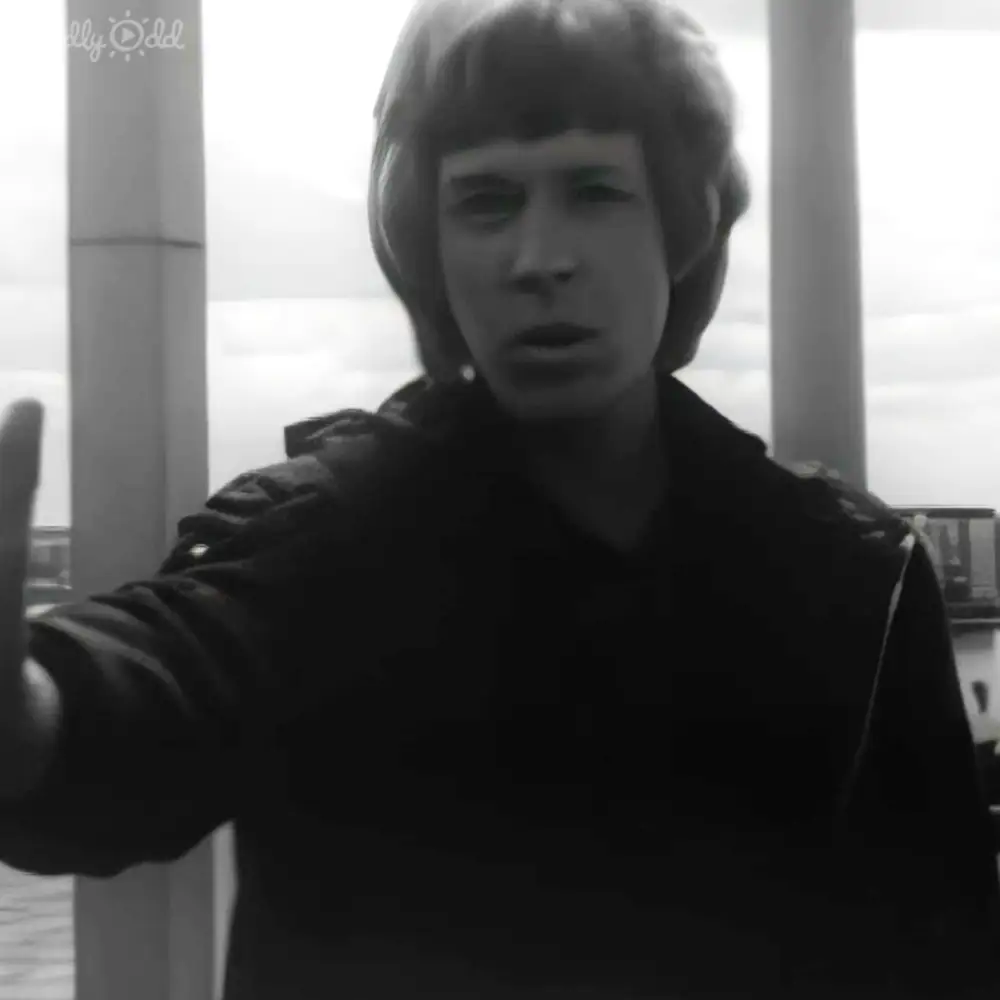 A young adult male in a dark-colored, structured jacket with shoulder pads and high collar stands outdoors against large pillars and water under cloudy skies.