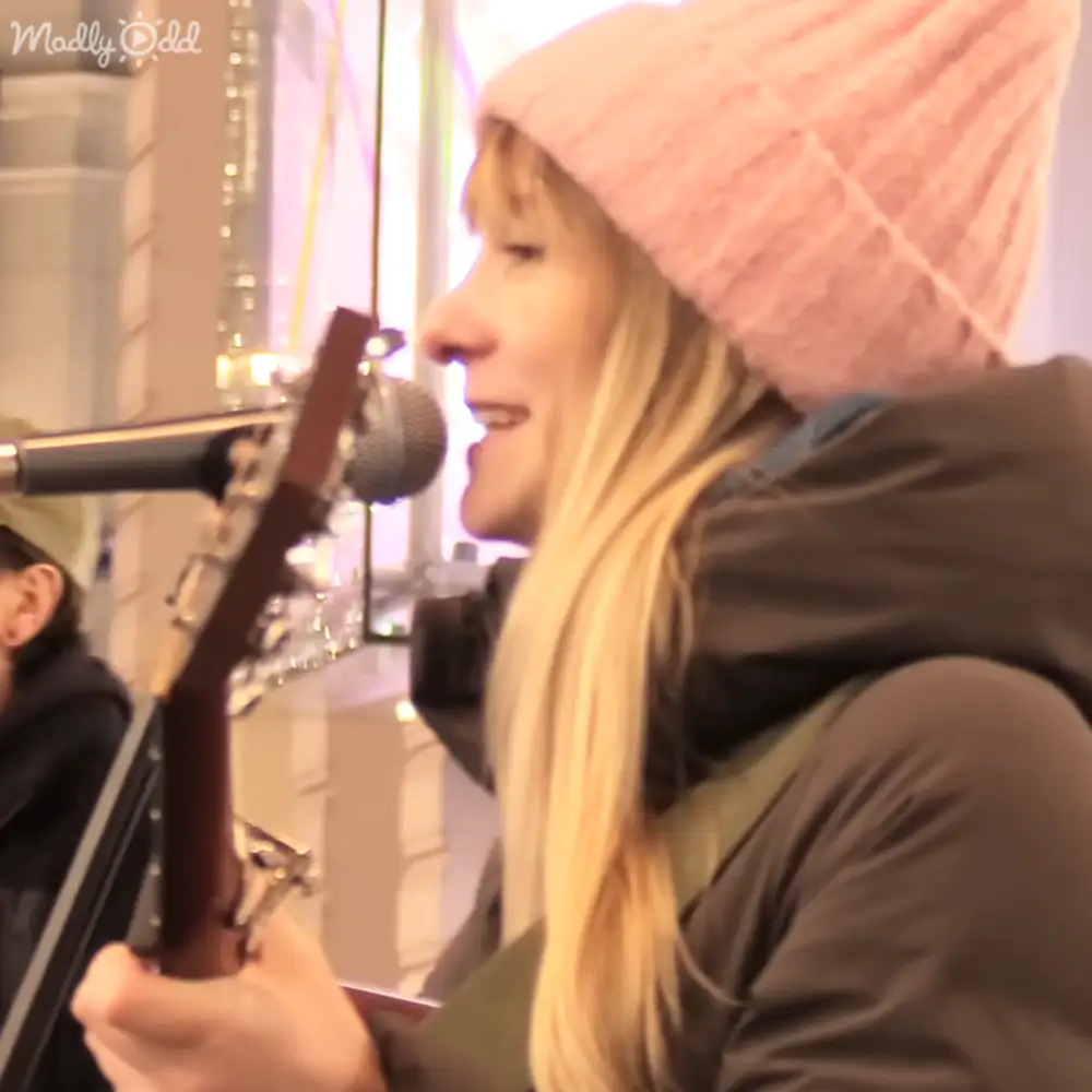 A young adult female wearing a pink knit beanie and dark green or black hooded jacket plays guitar in an indoor setting with blurred background elements suggesting a public area.