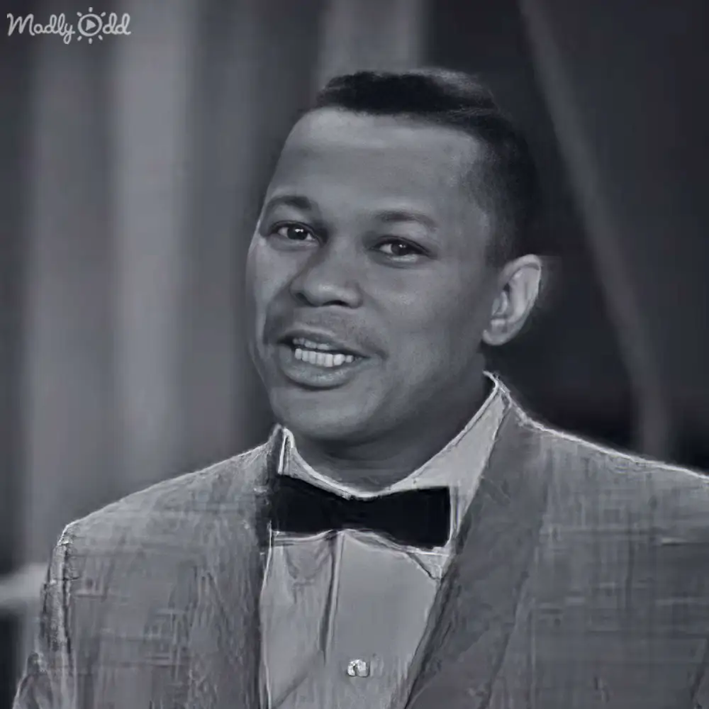 Adult male singer wearing a patterned suit jacket with a bow tie indoors against an elegant backdrop, possibly for a formal event or stage performance in black and white.