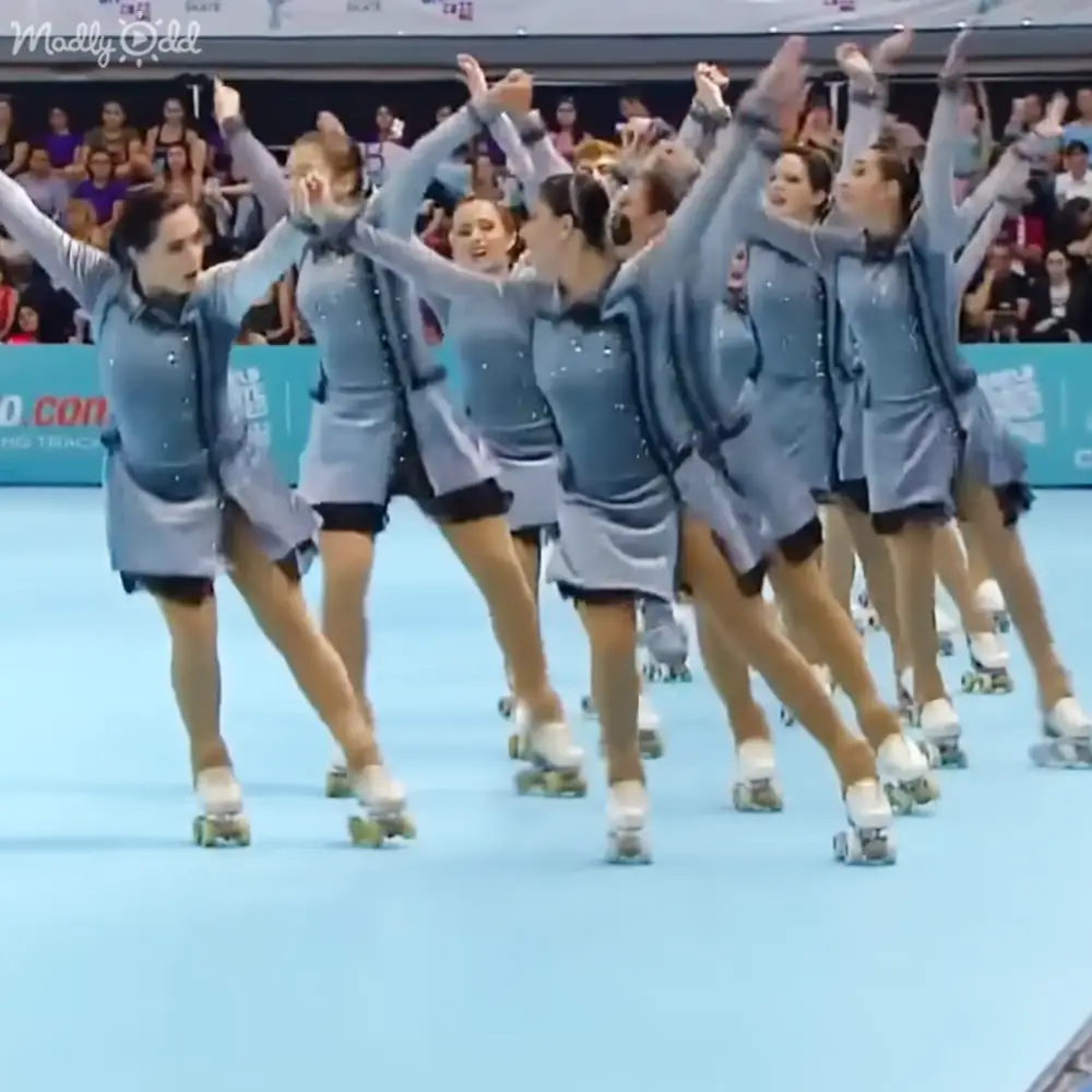 A group of young adult to middle-aged female roller skaters in light blue dresses perform indoors, with spectators seated and banners visible suggesting a competition or event.