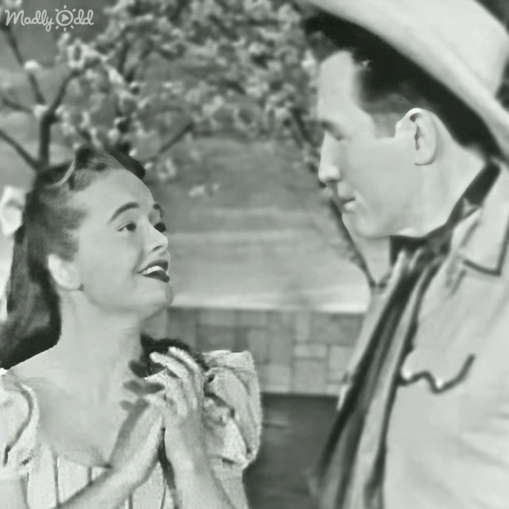 A young adult female and male stand outdoors, she wears a light-colored ruffled dress from mid-20th century, he wears a light jacket with darker lapels. The setting is an outdoor area with brick walls and greenery in bloom, suggesting springtime.