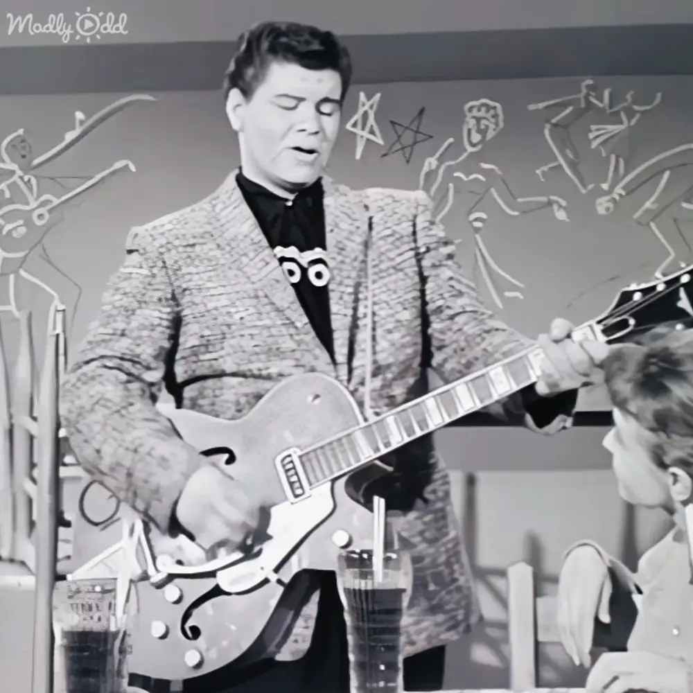 An adult male performer playing a guitar in an indoor studio setting with abstract line drawings on the wall and mid-20th-century fashion.