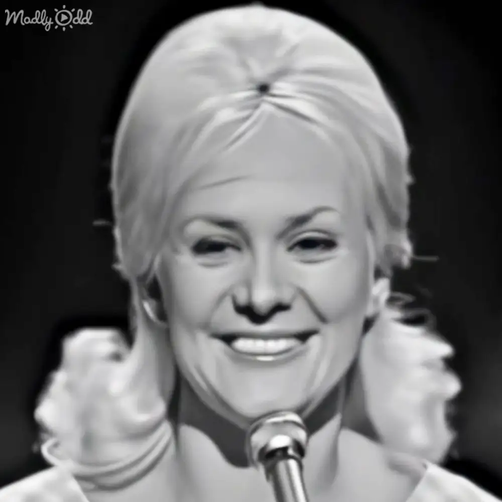 A middle-aged woman wearing a textured mid-20th-century blouse with a high collar stands indoors under dark lighting, possibly on stage.