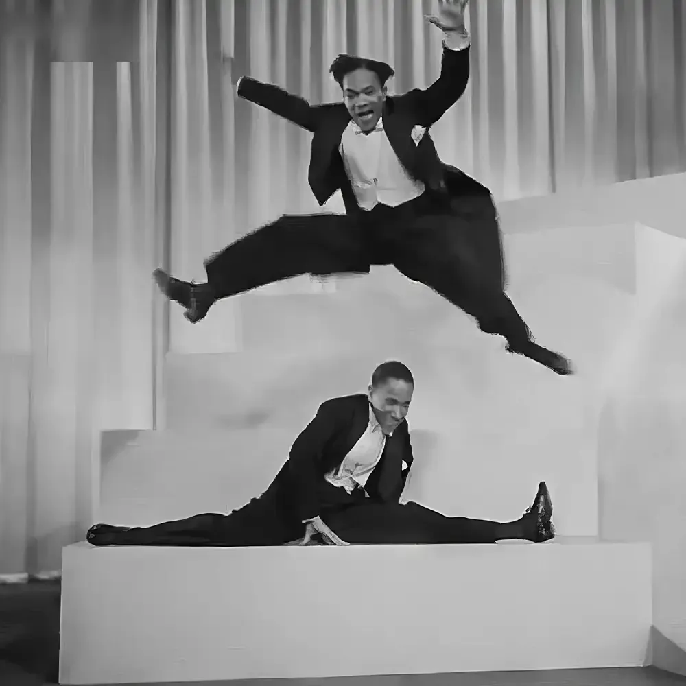 Two men dressed formally in dark suits with distinct styles pose on a stage with simple light-colored curtains as the backdrop.