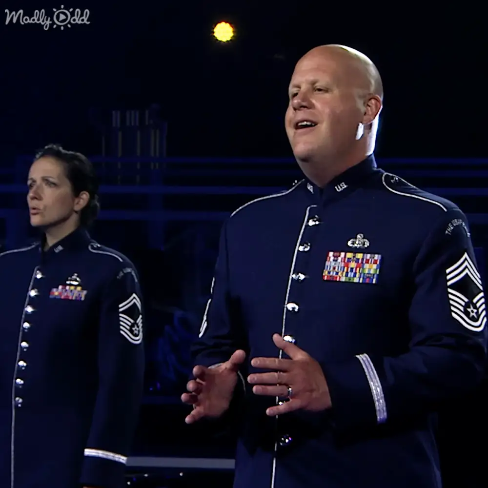 Adult male and female wearing navy blue military dress uniforms with stage lighting equipment visible in an indoor event setting under artificial light.