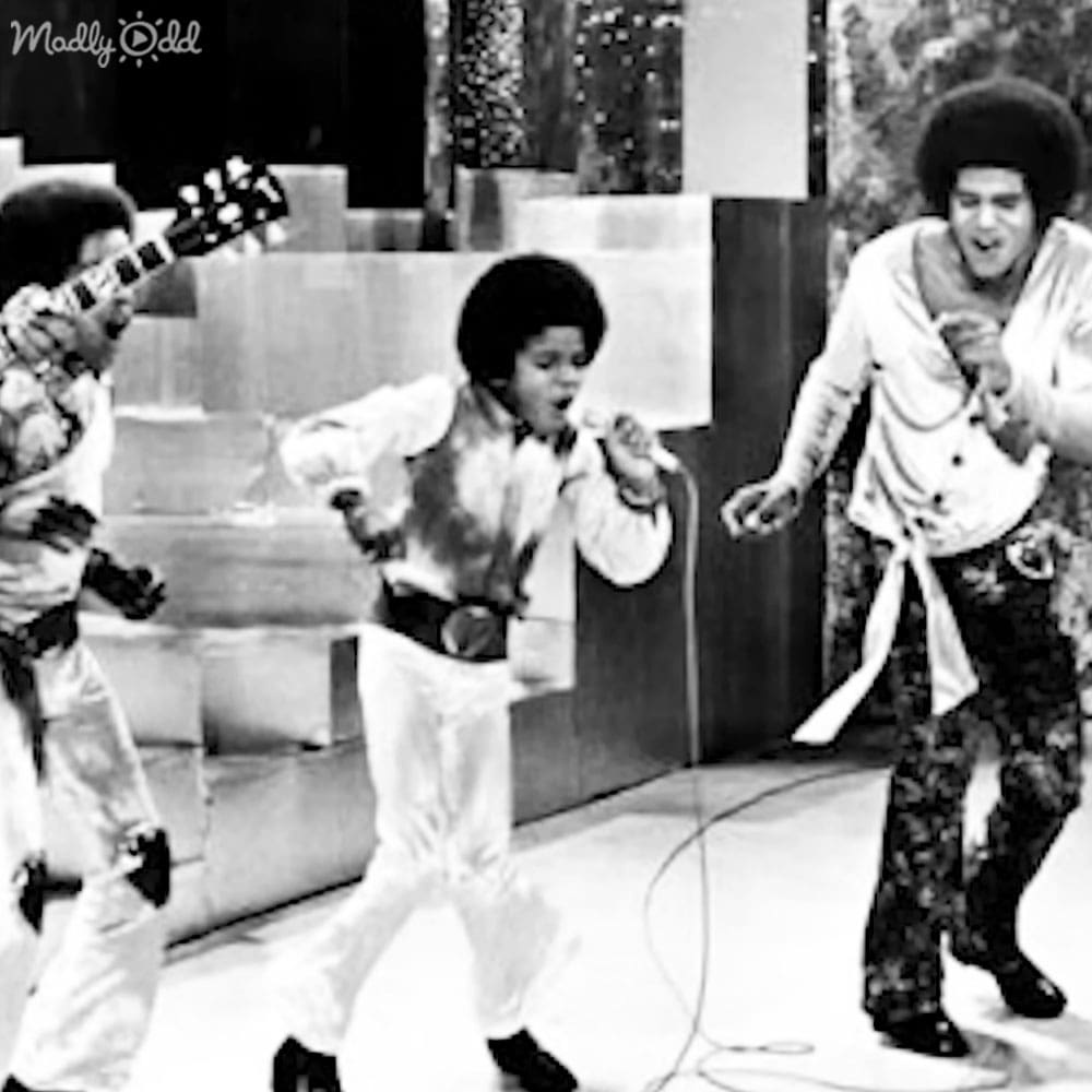 A young male performer wearing a 1970s-style white outfit with vest and pants, alongside adult males in similar attire, set against an indoor music performance backdrop.