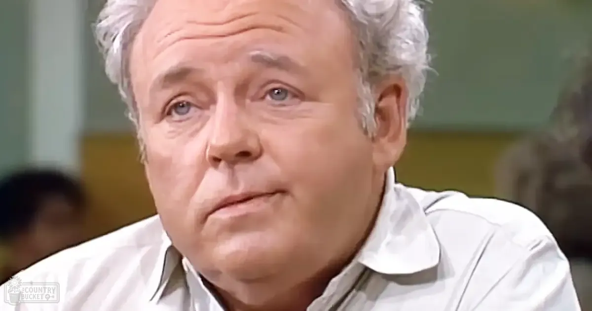 An older adult male in a casual white collared shirt, possibly mid-20th century style, is seated in an indoor setting with muted tones and soft lighting.
