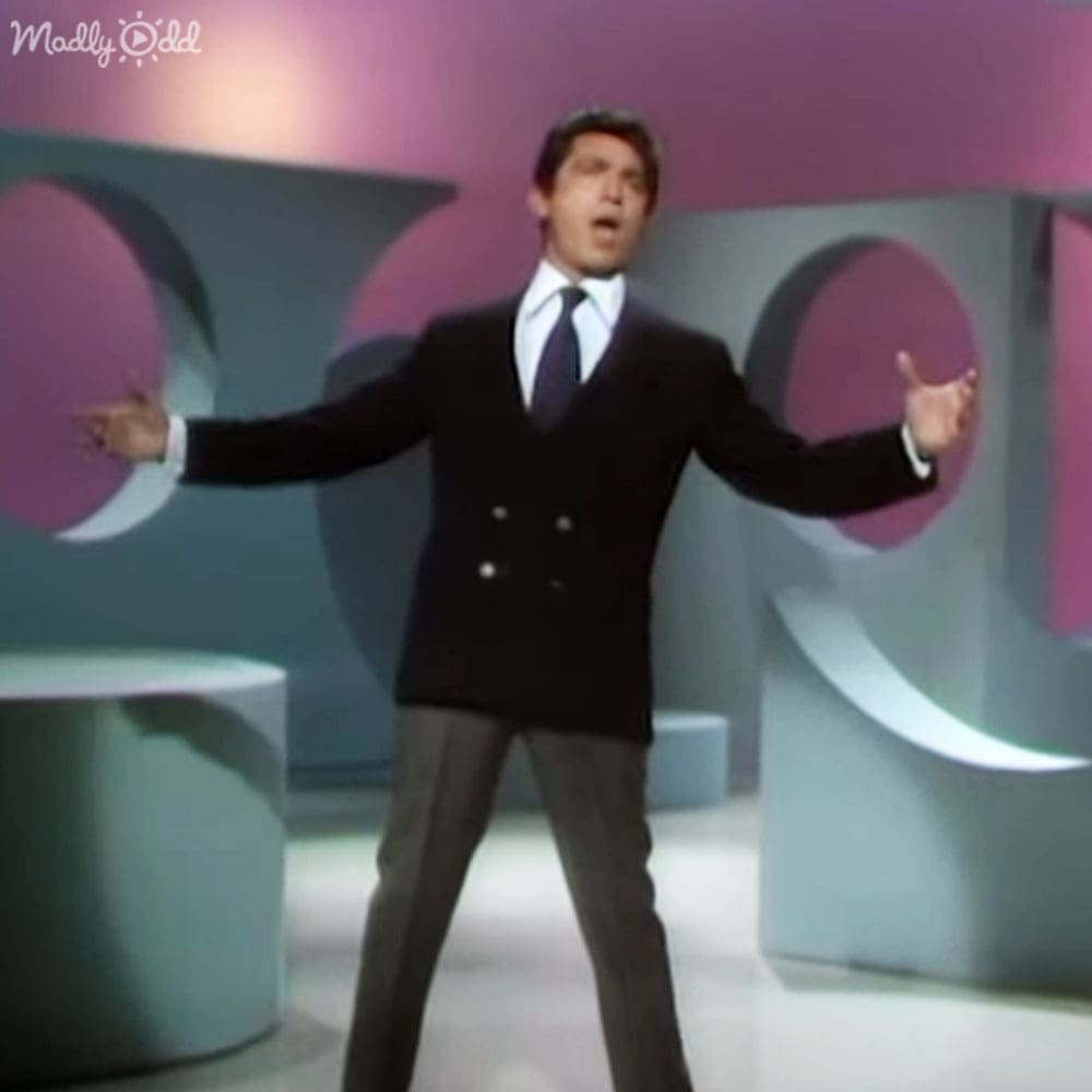 An adult male wearing a dark double-breasted suit jacket and white shirt stands indoors against an abstract, pastel-colored backdrop suggesting a 1960s or early 1970s television show set design.