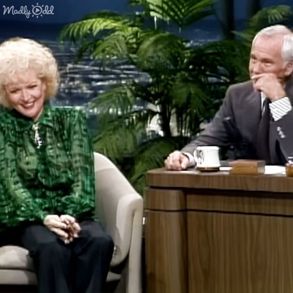 Two individuals in a talk show setting with greenery and indoor lighting, older adult female wearing a textured green patterned blouse and older adult male in a formal dark suit jacket and white checkered shirt, furniture includes a cushioned white armchair and wooden table with papers.