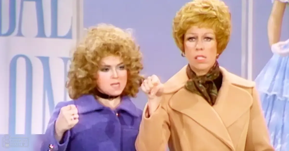 An adult female in a beige trench coat and scarf stands indoors with another adult female in a blue jacket against a backdrop suggesting a television studio setting.