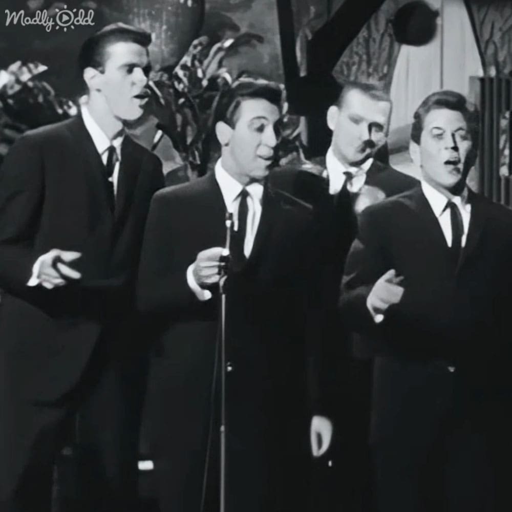 A mid-20th century musical performance group dressed in dark suits, standing on an indoor stage with decorative curtains and foliage.