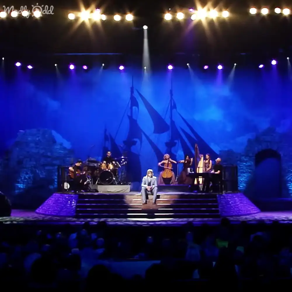 A musical performance in an indoor venue with a middle-aged male singer wearing a light-colored suit, accompanied by young and middle-aged musicians playing violins and drums; backdrop resembling ship's sails on water with evening lighting.