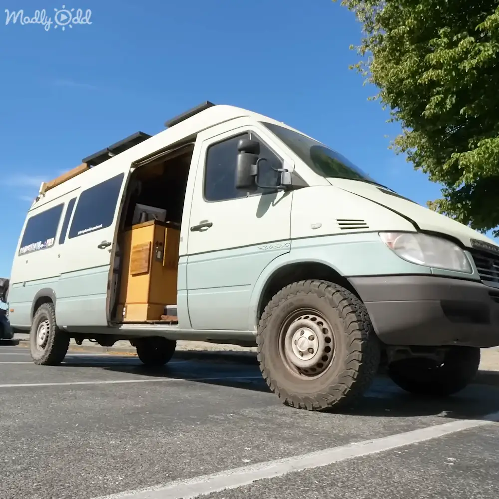 A white Mercedes-Benz Sprinter camper van with large off-road tires, parked in an outdoor parking lot surrounded by trees under a clear blue sky.