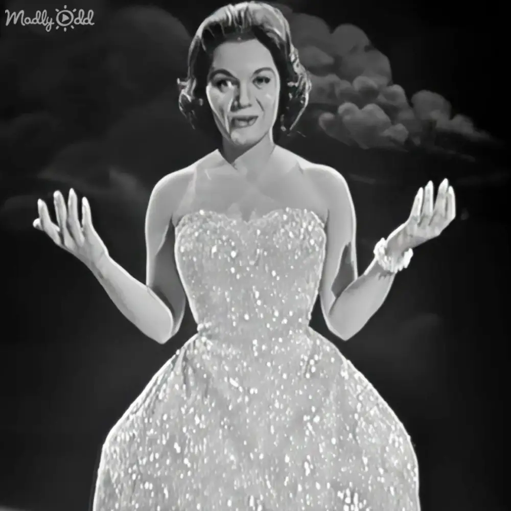 Adult female in an elegant strapless evening gown with a fitted bodice, full skirt, halter-style neckline, short sleeves, white or light-colored fabric, sparkly texture, and satin or silk material. Dramatic stage lighting emphasizes the attire against a backdrop of dark cloud-like shapes.