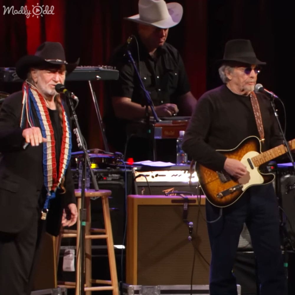 Musical performance at an indoor venue with performers wearing cowboy hats and formal attire, stage lighting focused on them against red curtains.