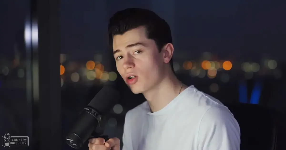 A young adult male in a white t-shirt holds a microphone, wearing a thin chain necklace. The setting is indoors at night with urban city lights visible.