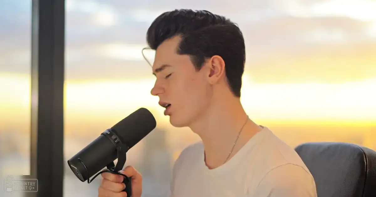A young adult male singer wearing a simple white t-shirt, holding a black microphone in an indoor setting with scenic outdoor views, suggesting early morning or late evening.