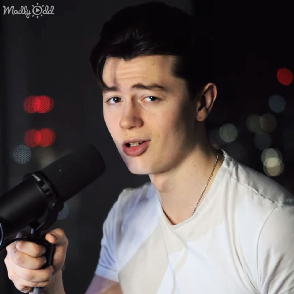 A young adult male wearing a white relaxed-fit t-shirt with a crew neckline, holding a black microphone in an indoor setting illuminated by artificial lighting.