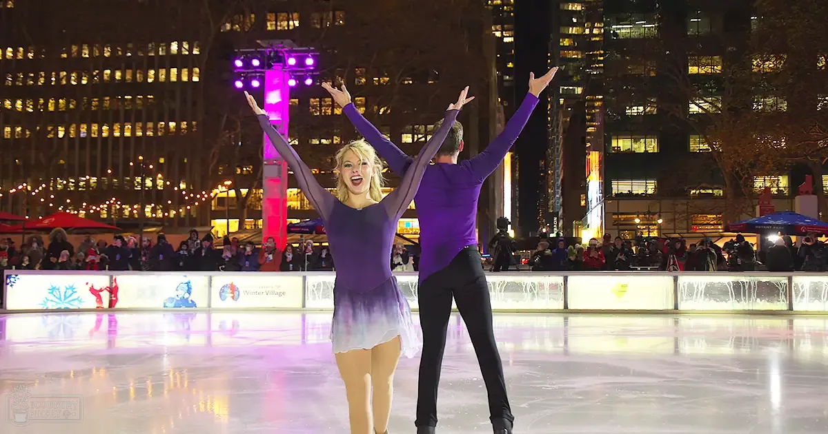 A couple ice skating at an outdoor rink under illuminated winter lights, surrounded by a crowd of spectators during what appears to be a public festival.
