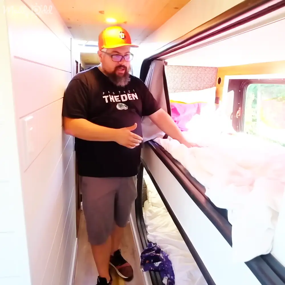 An adult male singer wearing a 'Protect The Den' T-shirt, gray shorts, and brown sneakers inside an RV with natural light during daytime. A purple cloth or garment lies on the floor near a bed covered in white bedding.