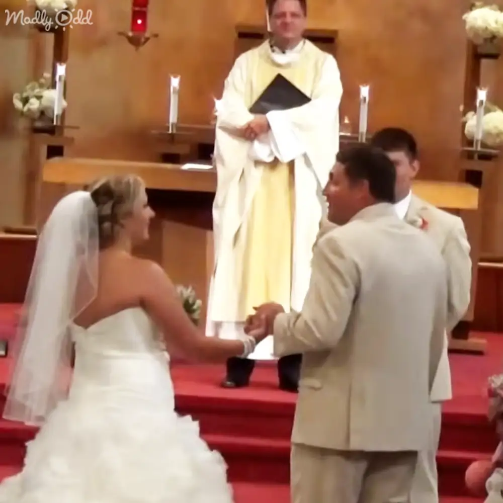 A wedding ceremony inside a church with red carpeted steps, white flowers, beige suits, and natural daylight.