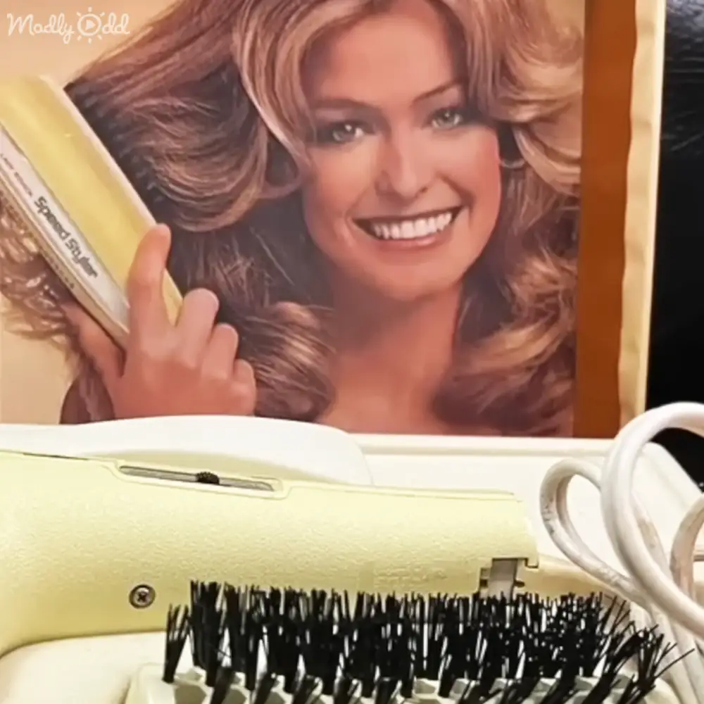 An adult female wearing a vintage 1970s-style garment with long sleeves and high neckline uses a Speed Styler hair dryer in an indoor setting, possibly a bathroom or bedroom.