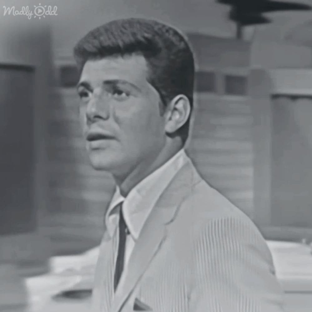 A young adult male in a light-colored suit jacket with vertical ribbed detailing, white shirt, and dark tie stands indoors.