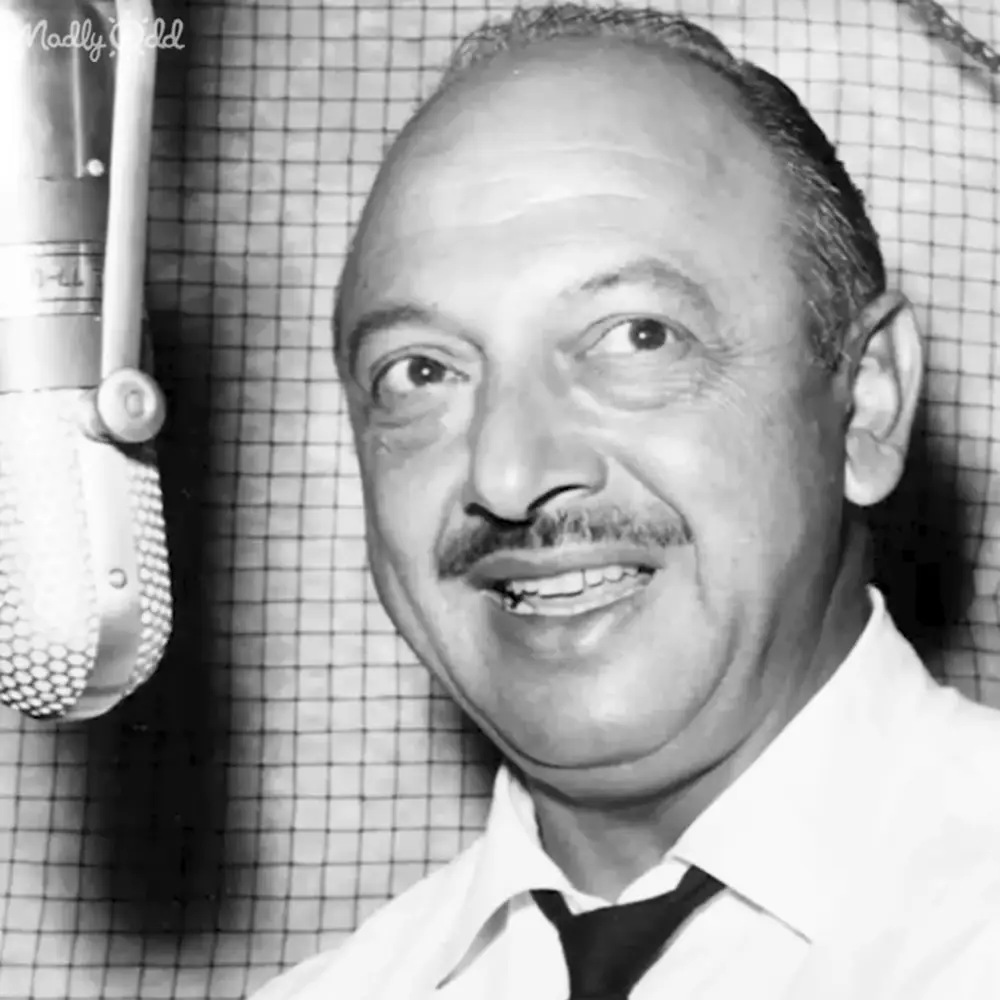 A middle-aged adult male in a formal white shirt with a black tie stands indoors near an older microphone, suggesting a recording studio environment.