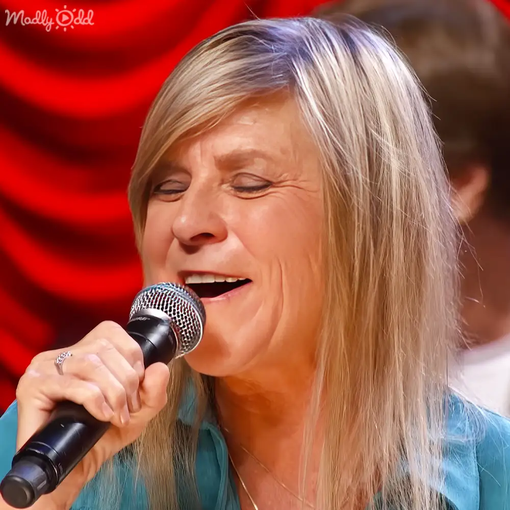 A middle-aged woman in a teal V-neck blouse sings into a microphone against a backdrop of red curtains, suggesting an evening stage setting.