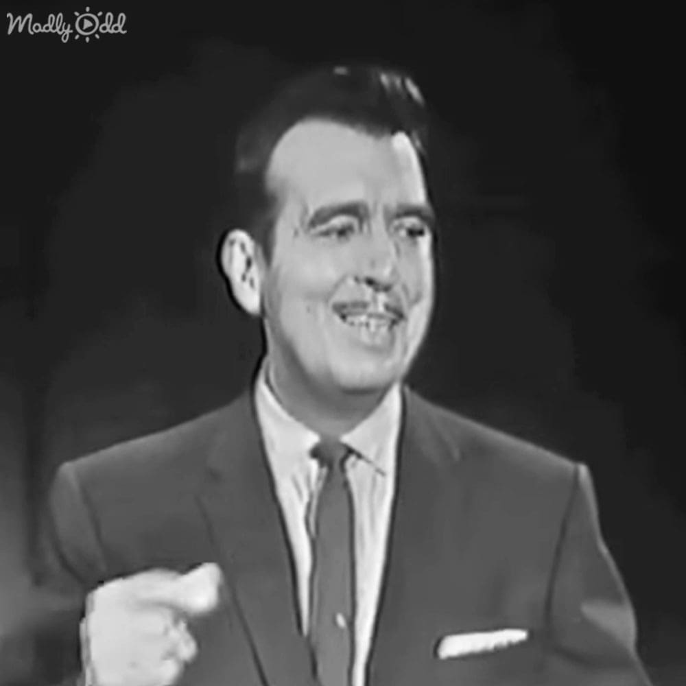 A middle-aged man in a dark suit jacket, white pocket square, light-colored shirt, and neatly knotted dark gray or black tie stands against a dark background with focused lighting typical of mid-20th century television shows.
