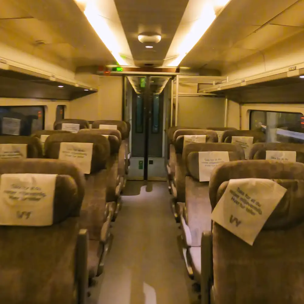 Rows of upholstered brown train seats with white coverings, overhead lights, door at center back, nighttime or early morning artificial lighting inside a train car.