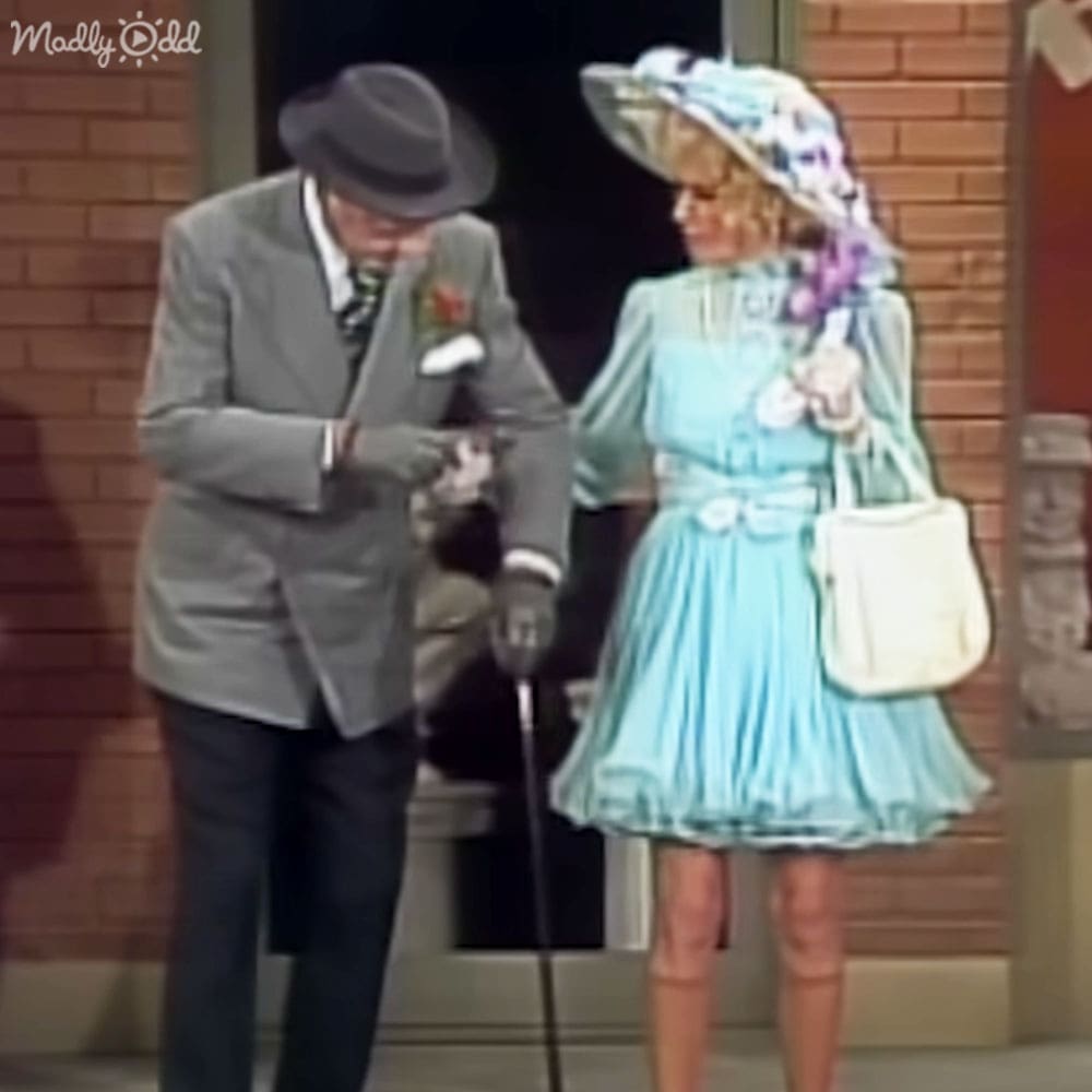 An older adult male singer wearing a gray suit jacket with a white pocket square and red flower, and an older adult woman in a light blue knee-length dress with a wide-brimmed hat adorned with flowers, set against a backdrop of a brick wall and stage elements under even lighting.