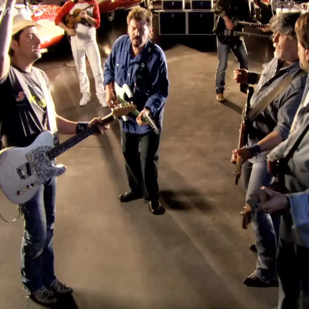A middle-aged man playing guitar in a garage workshop with another adult male holding a white electric guitar and two others in the background, one with a ukulele.