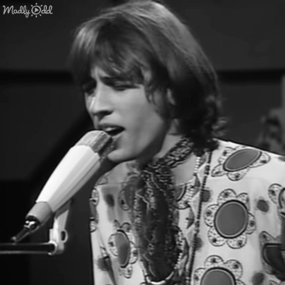 A young adult male sings into a microphone at an indoor event with dark curtains in the background.
