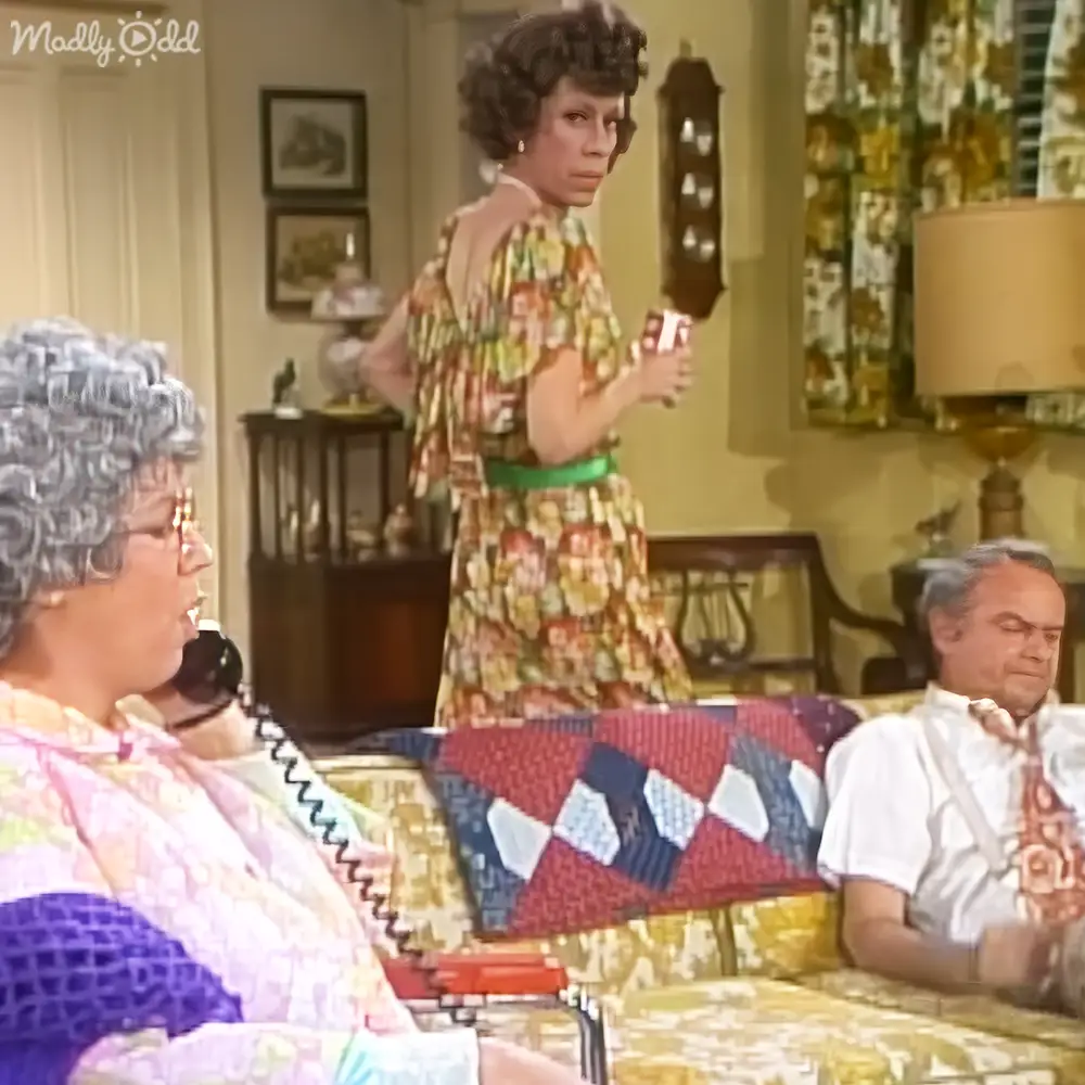 An adult female in a floral dress holds a can while seated with an older woman and another middle-aged man indoors, surrounded by furniture and home decor.