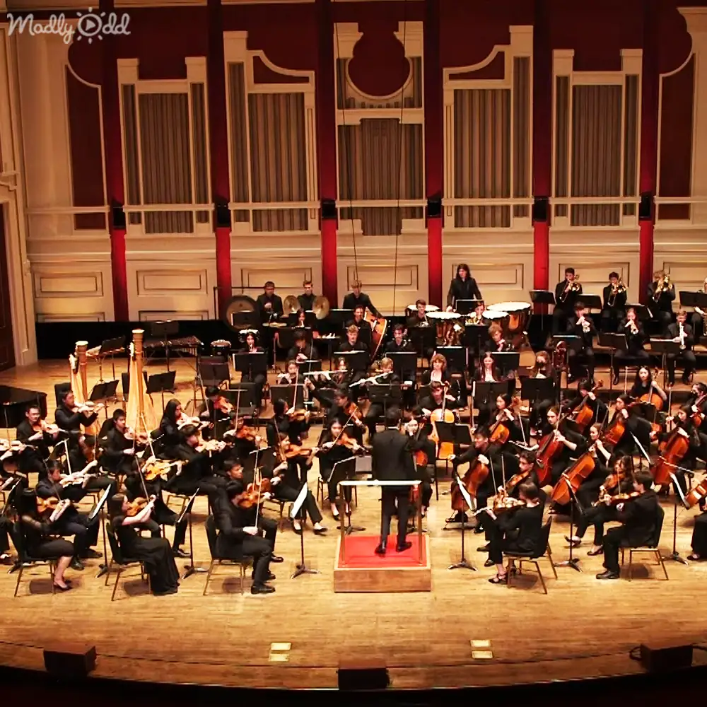 An adult male conductor leads a seated orchestra in formal attire within an ornate concert hall, with warm lighting highlighting the musicians under evening conditions.