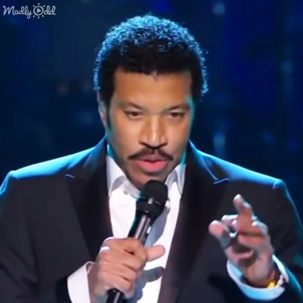 A male adult singer wearing a dark suit and holding a microphone on an indoor stage or performance venue.