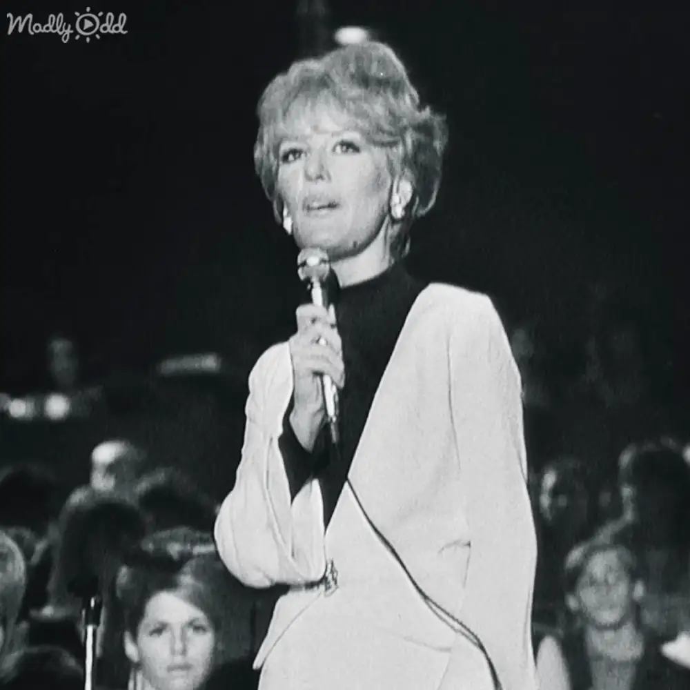 A middle-aged female performer wearing a white blazer with black turtleneck, standing on stage in an indoor concert setting.