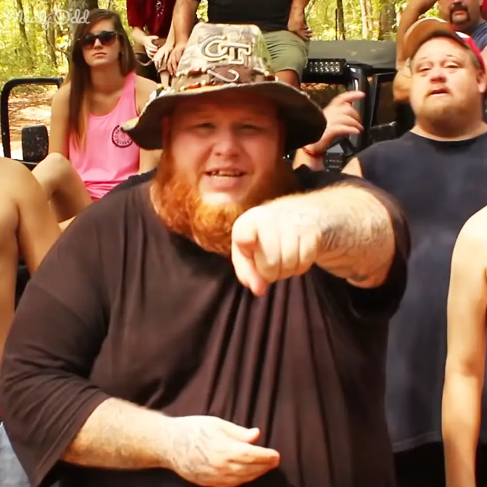 Adult male with red hair and beard wearing a hat, pointing forward outdoors amidst green foliage and other individuals in casual attire.