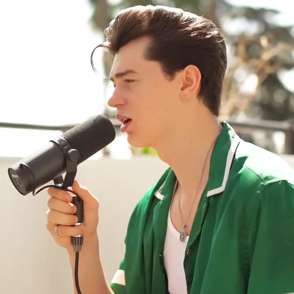 A young adult male wearing a green jacket with white trim and a white shirt, holding a microphone outdoors in an urban setting.