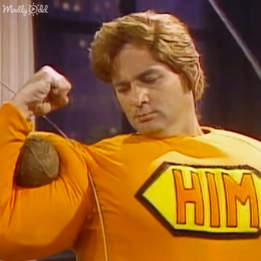 An adult male wearing an orange long-sleeved shirt with a bold yellow diamond-shaped patch that says "HIM", set indoors under artificial lighting.